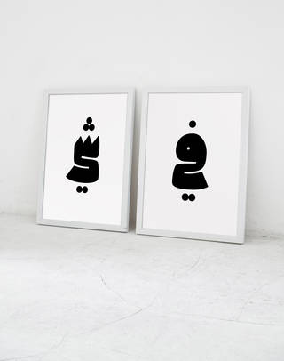 Two white-framed prints with black symbols on a white background, propped against a white wall on the ground
