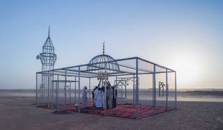 Chicken wire mosque construction in a sandy landscape against a blue sky with red carpet and worshippers inside