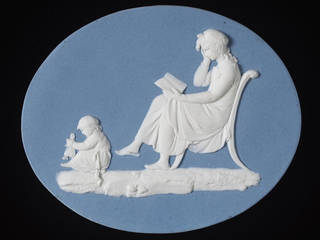 Flat oval plaque of white Jasperware with blue dip and applied white bas-relief decoration. A woman sits cross-legged on a chair in left profile, reading a book. She wears a mob-cap and holds her right hand up to her face. A child on the floor before her plays with a doll.