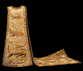 Brocade tabard, the floor-length front panel decorated with three heraldic lions, the back panel forming a train decorated all over with a diamond pattern. Appliqued with gold mesh, glass 'jewels', gold raffia, felt and imitation leather, coins and bottle tops, highlighted with gold paint. Lined with gold lamé, the lining of the train protected by a sheet of polythene.