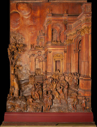 Detailed wooden carved panel in browns and orange tones with a crowd scene and the architural facades of buildings