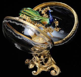 Clear crystal egg, shown open, on a gold stand, with a peacock on a branch inside