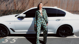 Person leaning against a white car, wearing a turquoise, green and cream patterned suit