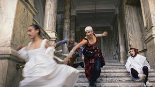 Person running down a grand staircase wearing a red ensemble. There is a person wearing white either side of them.