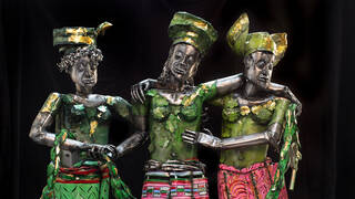 Three metal figures, wearing green and gold gèlè head ties. The figure representing Europe wears a Mondrian-like pattern; the figure representing Africa wears a West African Kente cloth and the  figure representing America wears Paisley.