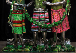 The legs of three metal figures. The figure representing Europe wears a Mondrian-like pattern; the figure representing Africa wears a West African Kente cloth and the  figure representing America wears Paisley. Draped across them is a wreath with a petrol nozzle at each end.