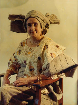 Photo of woman in traditional African fashion with head wrap, sat in chair