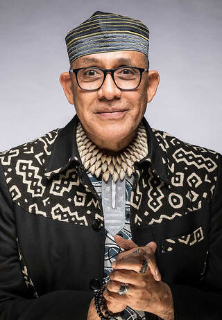 Portrait of Alphadi – a smiling man wearing black glasses, a shallow, rounded, striped hat, a layered shell necklace, rings on his fingers, and a black jacket with white patterned detailing around the shoulders.