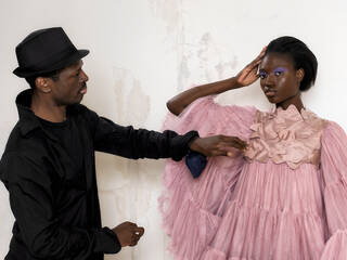 Man wearing black hat and black shirt, styling a model wearing a light pink voluminous dress