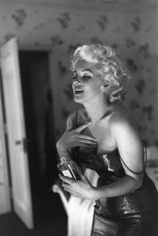 Woman with short, blonde wavy hair, holding Chanel perfume and applying it to her chest