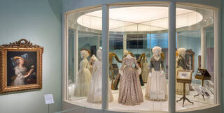 Round display case with a selection of period dresses in it and a portrait on the wall outside
