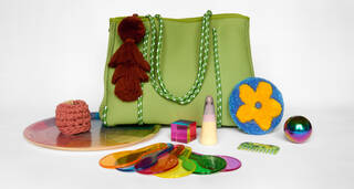 A green bag surrounded by colourful tacticle objects including a felted flower, colourful plastic viewfinders, a brown tassle and a small ceramic ghost 