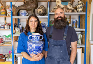 photograph of a man and a woman in blue overalls. The woman is holding a large blue and white pot covered in patterns and words