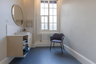 View inside the quiet room, looking towards the window. The walls of the room are painted off-white and the floor is blue linoleum. On the left wall there is a white washbasin with a large round mirror over it and a wooden storage cupboard built underneath. The storage cupboard has three shelves, holding prayer mats, blankets, and sensory mats. Opposite the basin, to the right, is a grey chair with armrests and four black legs. 