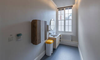 View inside the quiet room, looking towards the window. The walls of the room are painted off-white and the floor is blue and uncarpeted. Against the left wall there is a light switch dimmer, a baby-changing station, and a white waste-bin with a yellow lid. There is also a white washbasin with a large round mirror over it and a wooden storage cupboard built underneath. The storage cupboard has three shelves, holding prayer rugs, mat, and sensory toys. 