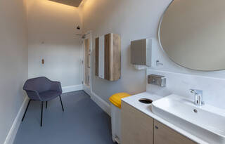 A view of the same room as before, now looking towards the door. There is a grey chair with arm rests and four black legs in the left corner. The chair slopes backward slightly. A Qibla is printed on the wall over the door. The door is manually operated by a pull handle.