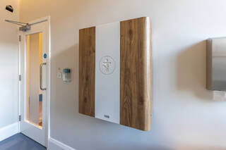 A wall-mounted baby changing table. The outer cover is faux-wood and white plastic. A tactile baby-changing pictograph is in the centre of the cover.