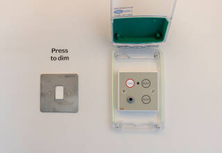 A white push button light switch mounted in a silvery metal plate. The words ‘press to dim’ are printed on the wall above the switch. Beside the switch, on the right, is the emergency call alarm, its protective case open so you can see the buttons operating the alarm. The buttons read ‘call’, ‘reset’, and ‘alert’.