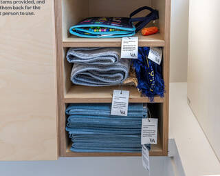 Close up of one of the wooden cupboards under the sink. The cupboards have three tiers of shelves holding sensory mat book and orange sensory toy, neatly folded blue and yellow prayer rugs with tassels, and folded blue mats.