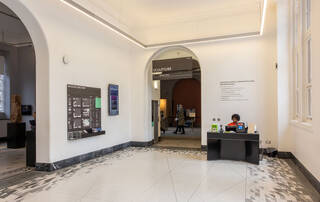 A large room with white walls and white marble flooring with black patterned edges. At the centre of the photo is an arched entry into the sculpture galleries. A sign hangs down from the entryway. The word ‘sculpture’ is printed in white lettering on a black background. To the left of the arched entry, a museum directory is mounted on the wall. There is also a screen with a digital display telling you what’s on at the museum. To the right of the arched entry is a small black desk with guidebooks, a computer, and hearing loops. A visitor experience assistant is seated at the desk wearing an orange museum uniform.