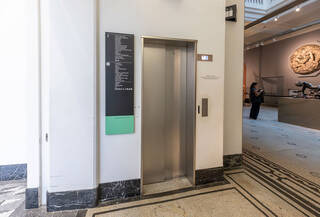 A lift with narrow, metal doors. The doors are closed. The floor indicator shows the left is coming down from floor 2, where the quiet space is. The walls surrounding the lift are white and a floor directory is mounted on the wall on the left side of the lift. Sculptures visible in the galleries to the right of the lift. The floor is black and white marble mosaic.