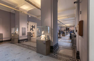 A long gallery with grey painted walls, high white ceilings, and white and black marble mosaic floors. A large square support column is front and centre of the photo. In front of it is a glass display case on a large plinth. Inside is a bust of a woman in white marble. Other sculptures mounted on plinths are visible down the centre and sides of the gallery.