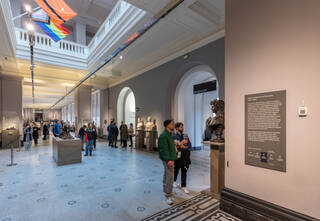A long gallery with grey painted walls, high white ceilings, and white and black marble mosaic floors. A large atrium is cut out of the floor above, adding to the spaciousness of the gallery. On the floor above are white parapet railings. The progress pride flag and refugee pride flags are mounted on flagpoles and hung from the atrium. There are visitors standing and walking through the hall, looking at sculptures mounted on plinths in the centre and along the walls of the room.