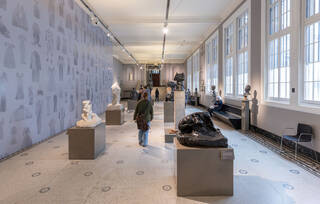 A long gallery with grey painted walls, high white ceilings, and white and black marble mosaic floors. Sculptures are displayed on plinths in two rows through the centre of the gallery and visitors are walking through and looking at the sculptures. The right walls of the gallery are lined with windows and bench seating. A temporary wall has been erected on the left side of the gallery, where the new fashion exhibit is being built. This wall is blue-grey and decorated with historical clothing items. 