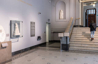 Corner of the Hintze sculpture gallery with doorway exiting to a lift corridor. The doorway is to the left of a white marble staircase with brass handrail. 