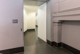 Doorway leading into the lift corridor. The corridor is wide enough to fit one wheelchair. The first part of the corridor is very short – about 1 wheelchair’s length – and then turns sharply to the right. The walls of the corridor are painted white and the floor is black.