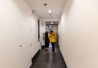  wide and can accommodate 16 standing people. The corridor is wide enough to fit one wheelchair. Two people, one in a black coat and trousers and another in a yellow coat and black trousers, are standing in front of the lift. The metal doors of the lift are opening. The corridor walls are painted white and the floor is black. The lift call buttons mounted on the wall on the left side. The lift directory is on the left adjoining wall.