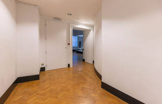 A wide corridor with white painted walls, black, high-contrast baseboards, and a wooden parquet floor. Bits of the corridor just out at slight angles. At the end of the corridor is a doorway entering to Design 1900-Now.