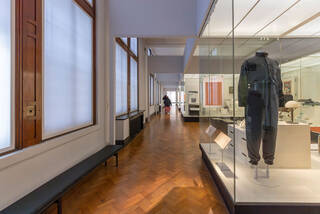 A long gallery with a series of large floor-to-ceiling glass cases on the right side of the room. The nearest case displays a headless mannequin wearing a dark grey jumpsuit. A row of shaded windows runs along the left side of the gallery. There are long black, backless benches under some of the windows. The corridor running between the cases and the windows is wide enough to accommodate a wheelchair. The floor is wood parquet. 