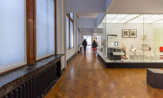 A view from the midpoint of the first Design 1900-Now gallery. On the left side of the wall under the windows is a long black radiator built into a black marble mantle. On the right side the gallery opens up into a wide area with mobile seating. The seats are black modular cubes without arms or backrests. Two visitors can be seen in the distance, walking through the gallery.