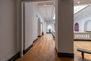 The atrium space between the two Design 1900-Now galleries. A large square support column dominates the right side of the picture. An arched doorway leading to the quiet space is on the left-hand side. A white parapet-style railing borders the atrium opposite the doorway.