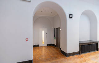 A large white arched doorway leading to a corridor with a lift on the right side and a closed white door at the end of the corridor. The flooring is wood parquet.